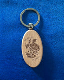 Das Produktbild zeigt einen ovalen Schlüsselanhänger aus Holz. Auf ihm ist das Bild eines Drachen, der auf einem Bücherstapel sitzt, sowie der Schriftzug "Im Herzen ein Drache" zu sehen.