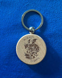 Das Produktbild zeigt einen runden Schlüsselanhänger aus Holz. Auf ihm ist das Bild eines Drachen, der auf einem Bücherstapel sitzt, sowie der Schriftzug "Im Herzen ein Drache" zu sehen.