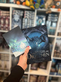 Auf dem Bild ist eine Hand abgebildet, welche das Buch "Der Spiegel des Drachen" und die dazugehörige Charakterkarte hält. Auf der Karte ist ein weißer, neunschwänziger Fuchs abgebildet, welcher vor einem See steht. Im abgebildeten Himmel kann man Polarlichter erkennen.