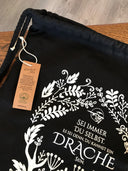 Das Produktbild zeigt einen schwarzen Turnbeutel.  Das Drachenmond-Logo (ein fliegender Drache vor einem Vollmond) ist mittig auf den Beutel gedruckt. Unter dem Logo steht "Sei immer du selbst, außer du kannst ein Drache sein". Unten am Beutel ist ein großer weißer Drache mit Hörnern abgebildet. Von diesen Hörnern aus ziehen sich verschiedene weiße Blätter und Ranken um das Logo und die Schrift herum.