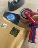 Auf dem Bild sieht man einen gepolsterten Umschlag mit grünem Absenderetikett, darüber einen Paketband-Abroller, einen Tesafilm-Abroller und eine Drachenmond-Postkarte. 
