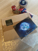 Auf dem Bild sieht man ein Päckchen mit grünem Absenderetikett auf einem Stück Luftpolsterfolie und darüber - etwas unscharf - einen Paketband-Abroller. Auf dem Päckchen sieht man die Drachenmond-Postkarte liegen. 