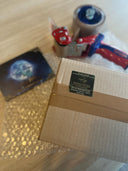 Auf dem Bild sieht man ein Päckchen mit grünem Absenderetikett auf einem Stück Luftpolsterfolie, darüber - etwas unscharf - einen Paketband-Abroller und eine Drachenmond-Postkarte. 