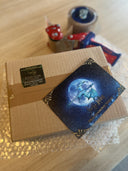 Auf dem Bild sieht man ein Päckchen mit grünem Absenderetikett auf einem Stück Luftpolsterfolie und darüber - etwas unscharf - einen Paketband-Abroller. Auf dem Päckchen sieht man die Drachenmond-Postkarte liegen. 