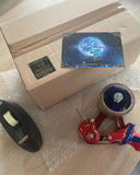 Auf dem Bild sieht man ein großes Paket mit grünem Absenderetikett auf einem Stück Luftpolsterfolie, davor einen Paketband-Abroller, einen Tesafilm-Abroller, darauf eine Drachenmond-Postkarte. 
