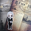 Das Magnetlesezeichen zeigt die Figur "Dietrich" des Buches "Ruf der Rusalka". Es ist im Anime-Stil gezeichnet und zeigt einen weißhaarigen Mann in einem schwarzen Frack. Darunter trägt er ein weißes Hemd mit weißer Fliege und über seinem angewinkelten linken Arm liegt ein weißes Serviertuch.