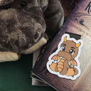 Das Produktbild zeigt ein Magnetlesezeichen mit einem Drachen in den Farben Braun und Beige. Es liegt auf einem kleinen Stapel Bücher. Im Hintergrund sitzt der Plüschdrache Djiwar.
