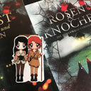 Das Produktbild zeigt die beiden Hauptfiguren aus dem Buch "Rosen & Knochen". Links sieht man Muireann, mit dunklem Haar, einer hellgrünen Weste über einem rötlichen Hemd, einer dunklen Hose und einem dunklen Miedergürtel, an dem ein Dolch und ein Beutel stecken. Rechts sieht man Rose mit roten Locken, einer braunen Tunika, brauner Hose, einem Dolch am Gürtel und einem Stab in der Hand. Das Lesezeichen liegt auf den dazu passenden Büchern. 