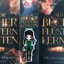 Das Magnetlesezeichen zeigt die Protagonistin des Buches "Als Bücher flüstern lernten". Es ist im Anime-Stil gezeichnet und zeigt eine dunkelhaarige Frau in einem grün-schwarzen altertümlichen Jägerinnenoutfit. Auf ihrem Rücken ist eine Axt geschnallt. Im Hintergrund sind die dazu passenden Bücher abgebildet.