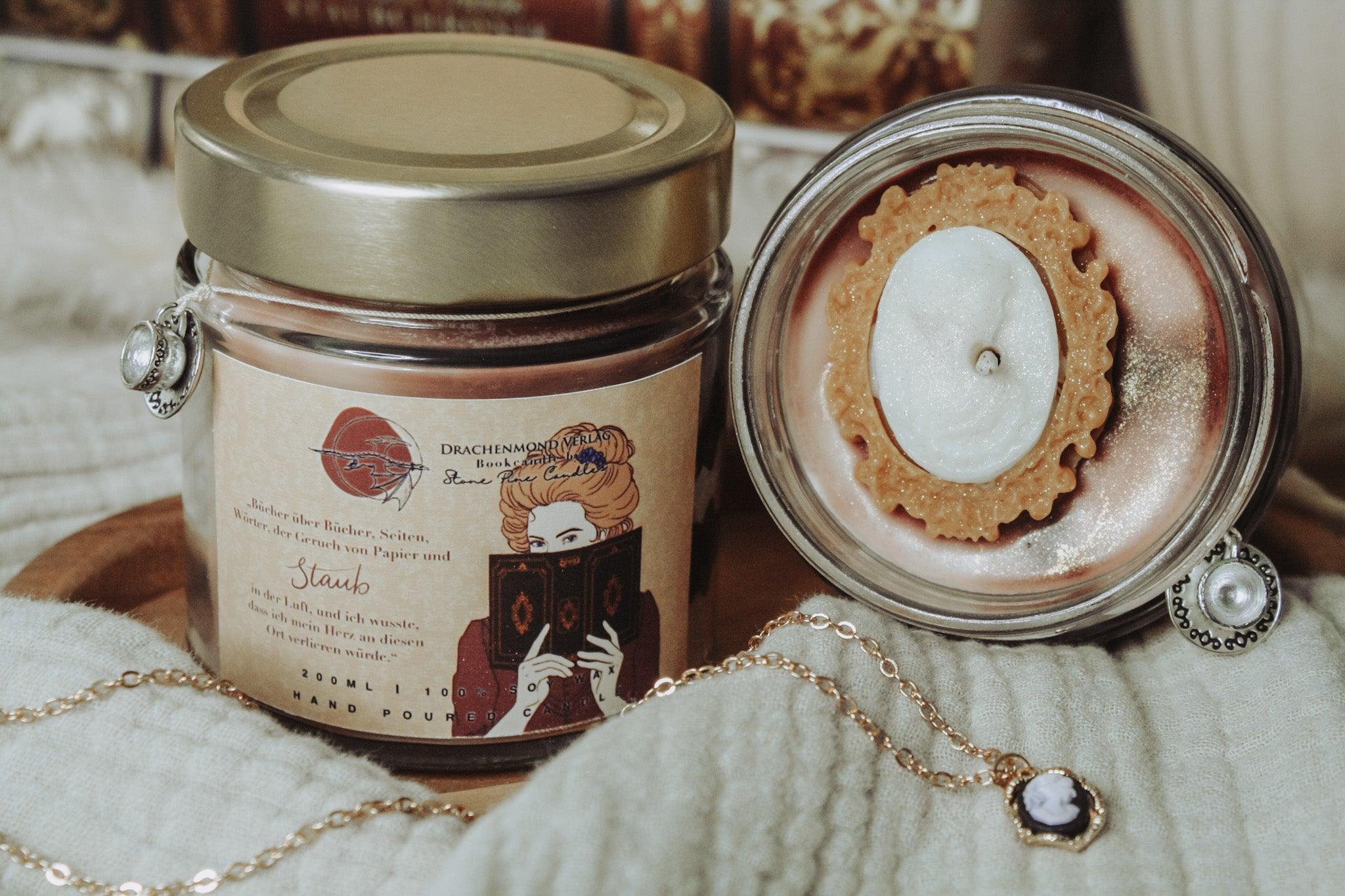 Das Produktbild zeigt zwei Kerzen zu Animant Crumbs Staubchronik. Auf dem Label der linken Kerze ist eine Frau abgebildet, die den Betrachter über ein Buch hinweg anschaut, daneben steht ein Zitat: 