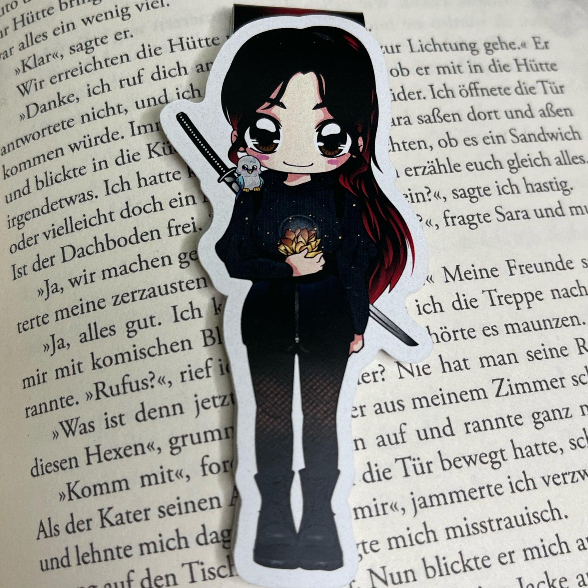Das Produktbild zeigt ein Magnetlesezeichen mit einer dunkelhaarigen Frau in schwarzem Oberteil und schwarzen Netzstrumpfhosen. Auf dem Rücken trägt sie einen Katana, auf ihrer Schulter sitzt ein Wellensittich und in der Hand hält sie eine goldene Lotusblüte. Das Lesezeichen liegt auf einer Buchseite. Es ist im Anime-Stil gezeichnet.