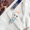 Das Produktbild zeigt ein Magnetlesezeichen mit einer blonden Frau. Sie trägt ein weißes Shirt, darüber einen langen grünen Cardigan, eine blaue Hose und orange-weiße Schuhe. In ihrer linken Hand hält Sie einen orangenen Becher und auf ihrer Schulter sitzt ein braun-weißer Hamster. Gezeichnet ist das Lesezeichen im Anime-Stil.