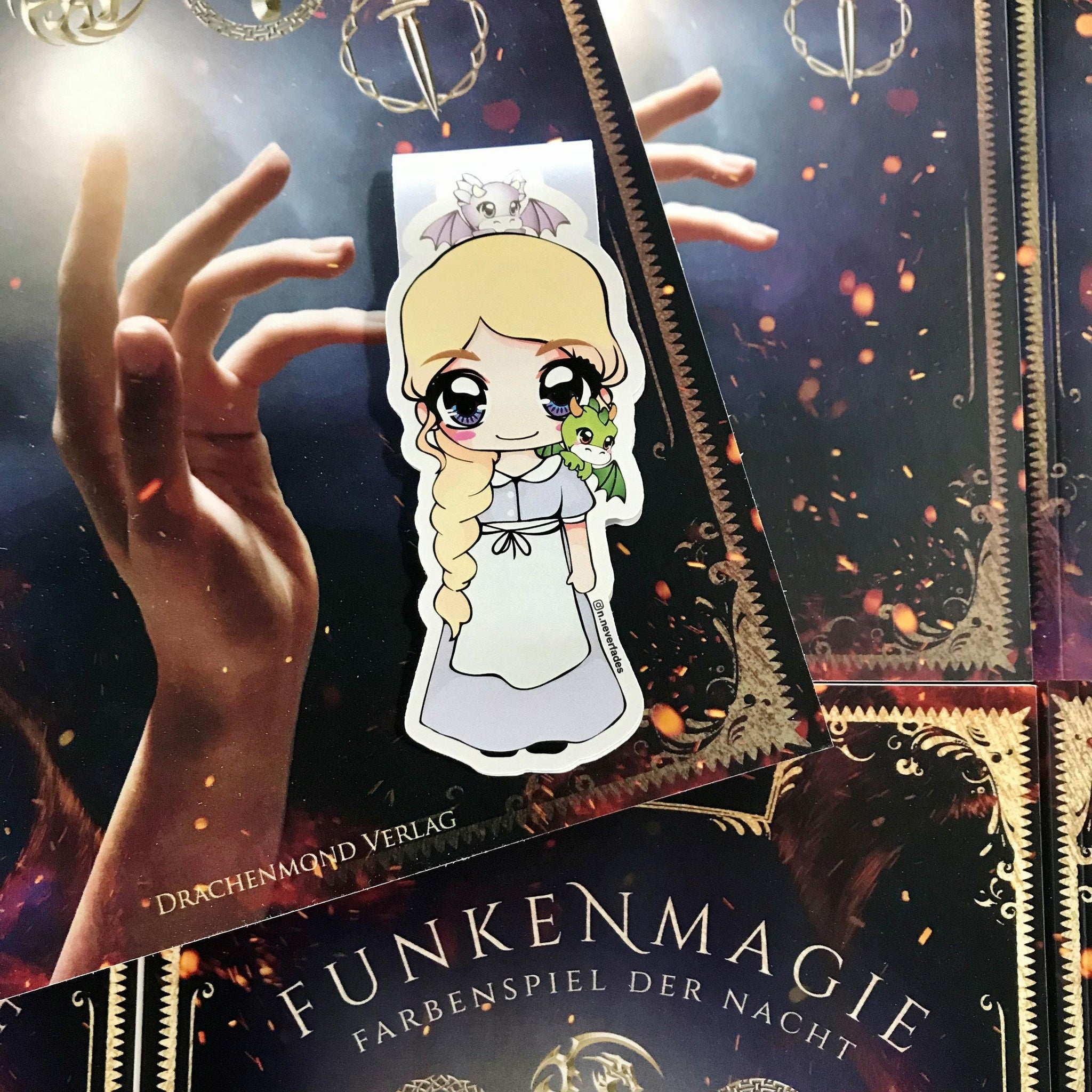 Das Produktbild zeigt ein Magnetlesezeichen mit einer blonden Frau in einem langen Dienstmädchenkleid. Auf ihrer Schulter sitzt ein kleiner grüner Drache, auf ihrem Kopf ein hellgrauer Drache. 