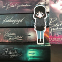 Das Magnetlesezeichen zeigt die Figur Meg aus der Buchreihe "Ein Roman über die Anderen". Es ist im Anime-Stil gezeichnet und zeigt eine junge Frau mit schulterlangen, dunklen Haaren. Sie trägt ein dunkelgraues Oberteil, einen kurzen schwarzen Rock und graublaue Hosen. In der Hand hält sie eine Klinge. Das Lesezeichen lehnt an einem Stapel der zugehörigen Reihe. Auf den Buchrücken kann man den jeweiligen Titel lesen. 