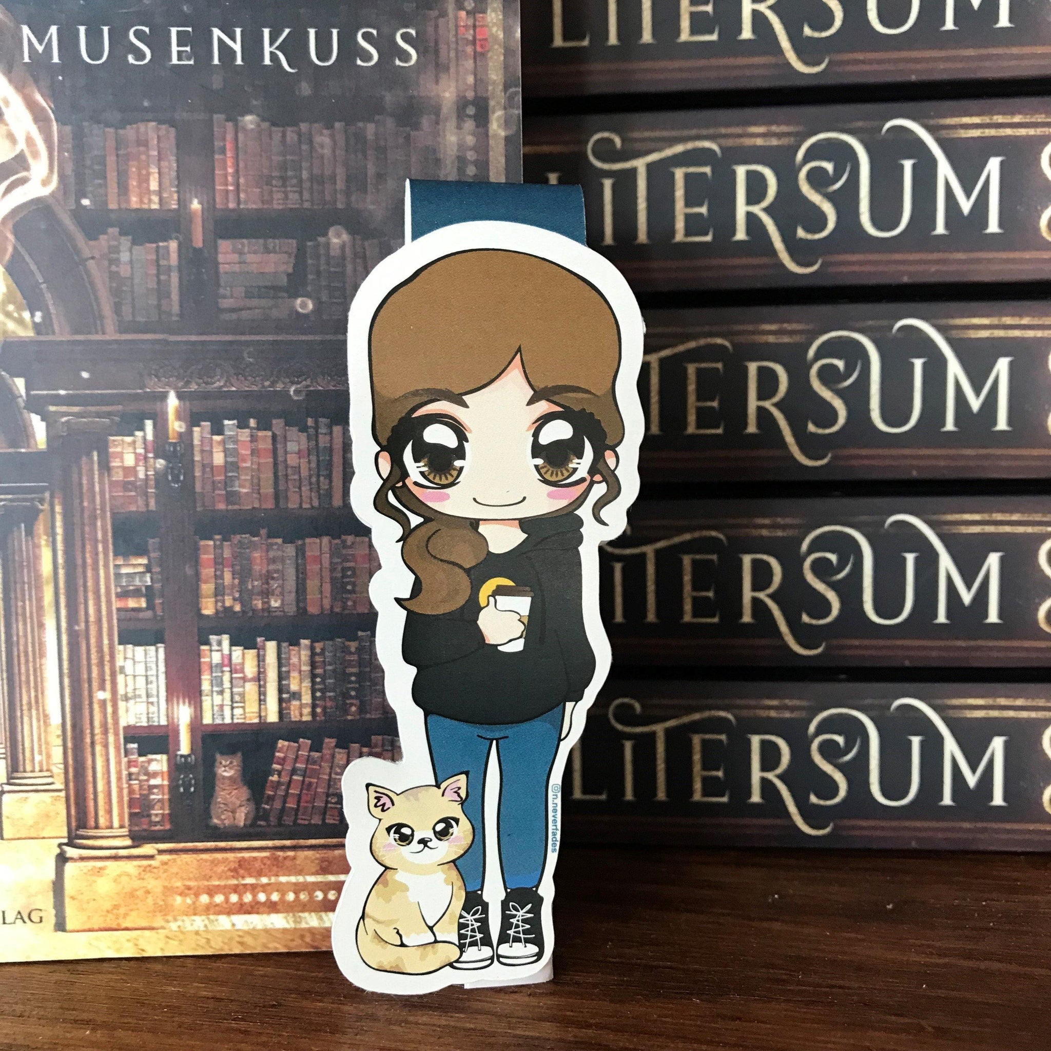 Das Produktbild zeigt ein Magnetlesezeichen mit einem brünetten Haaren. Sie trägt einen schwarzen Hoodie, eine blaue Hose und schwarze-weiße Schuhe. In ihrer rechten Hand hält sie einen Kaffeebecher und neben ihr sitzt eine weiß-beige-braune Katze. Gezeichnet ist das Lesezeichen im Anime-Stil. 