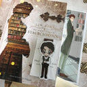 Das Produktbild zeigt ein Magnetlesezeichen vor dem dazu passenden Buch "Staubchronik". Auf dem Lesezeichen sieht man einen Mann mit kurzen dunklen Haaren und einer Lesebrille. Er trägt eine braune Hose, eine braune Weste und eine rote Fliege. Vor seiner Brust hält er ein Buch. Gezeichnet ist das Lesezeichen im Anime-Stil.