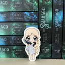 Das Magnetlesezeichen zeigt die Protagonistin des Buches "Der Smaragddrache". Die blonde Frau, deren geflochtener Zopf über ihrer linken Schulter bis zu ihren Knien reicht, trägt ein hellblaus Kleid, mit langen Ärmeln und keine Schuhe. Auf ihren Schultern schlängelt sich ein kleiner dunkler Drache. Das Lesezeichen ist im Anime-Stil gezeichnet.