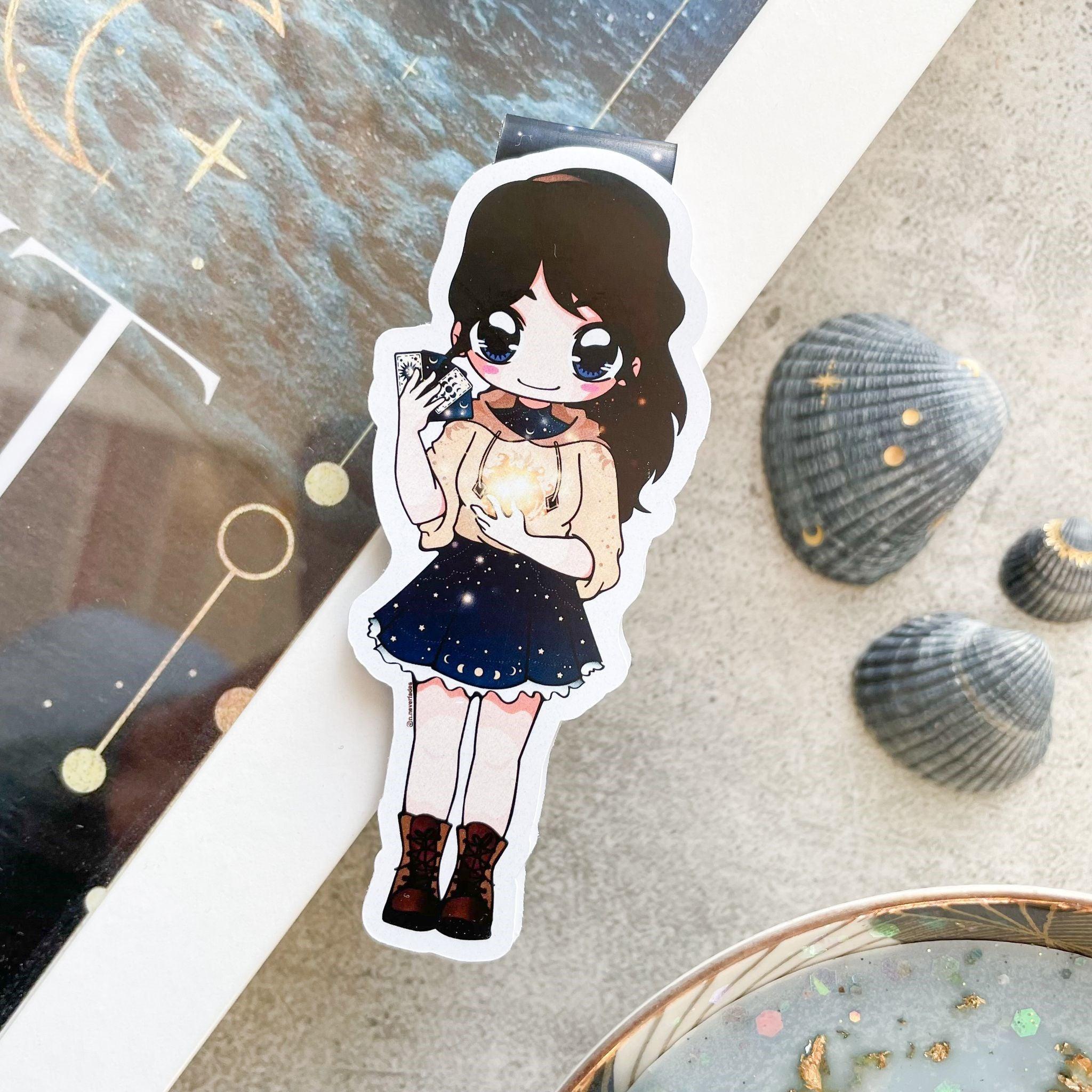 Das Produktbild zeigt ein Magnetlesezeichen brünetten Frau in einen dunkelblauen Kleid. Auf dem Kleid sind kleine Sterne und Monde abgebildet. Darüber trägt sie einen beigen Hoodie. In ihrer rechten Hand hält sie Tarotkarten. Gezeichnet ist das Lesezeichen im Anime-Stil.